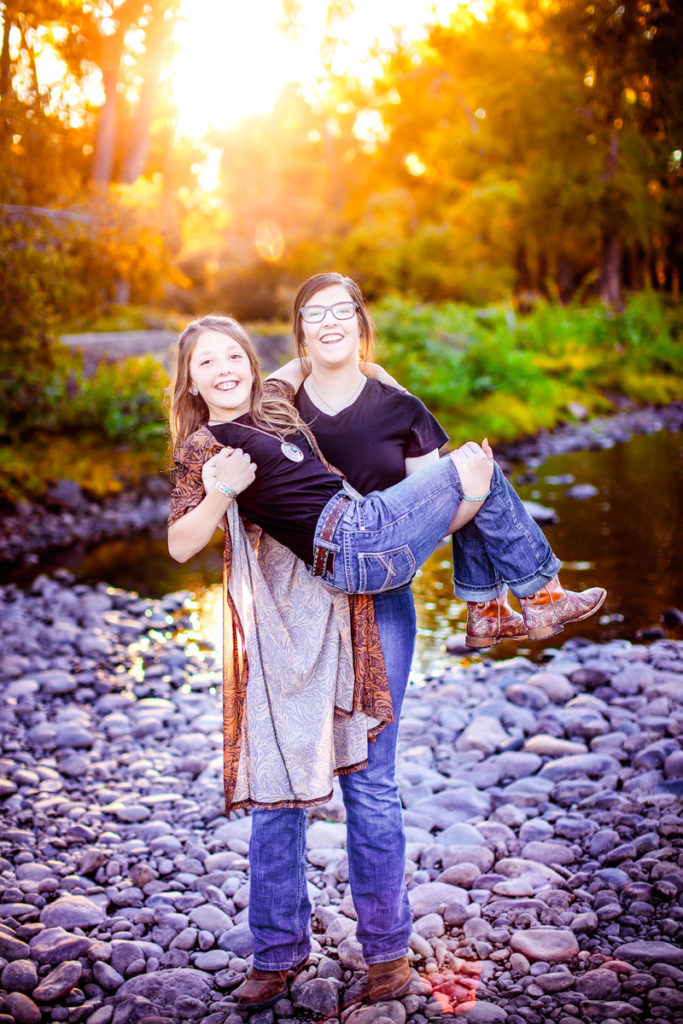 Family Photography, Teenage girl holds sister in her arms, both smile near a riverbed