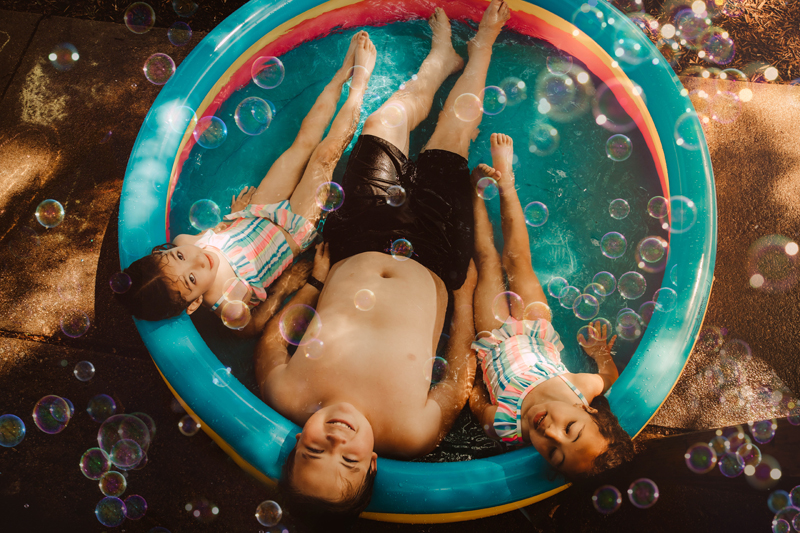 Family Photographer, Brother and two younger sisters relax and lay back in a kiddy pool in the grass
