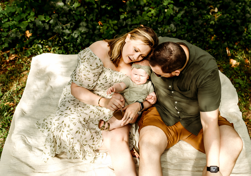Newborn Photography, Mom and dad hold baby, they sit on a picnic blanket outside